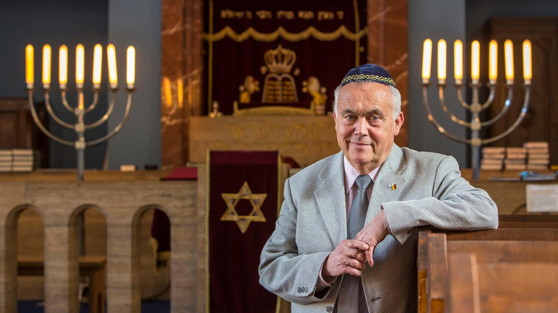 Reinhard Schramm, Vorsitzender der Jüdischen Landesgemeinde Thüringen, in der Synagoge in Erfurt. Schramm, der eine blaue Kippa trägt, hat seinen Arm und die Lehne einer Bank gelegt. Im Hintergrund spenden jüdische Leuchter Licht.