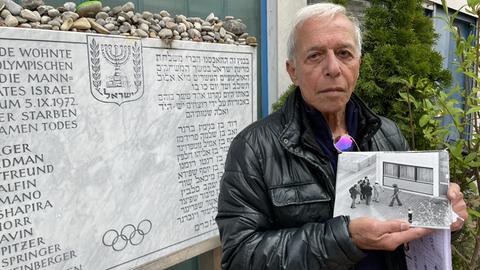 Shlomo Levy posiert mit einem historischen Foto neben der Gedenktafel im Olympischen Dorf in München für ein Foto.