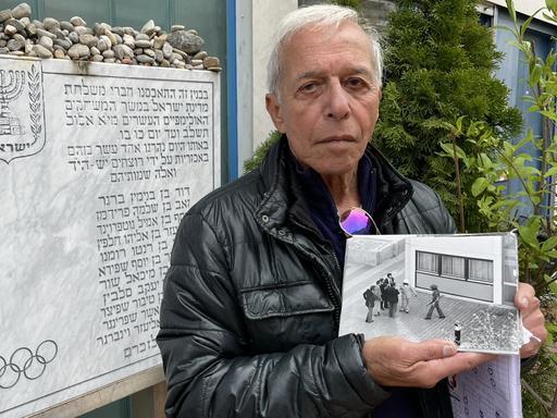 Shlomo Levy posiert mit einem historischen Foto neben der Gedenktafel im Olympischen Dorf in München für ein Foto.
