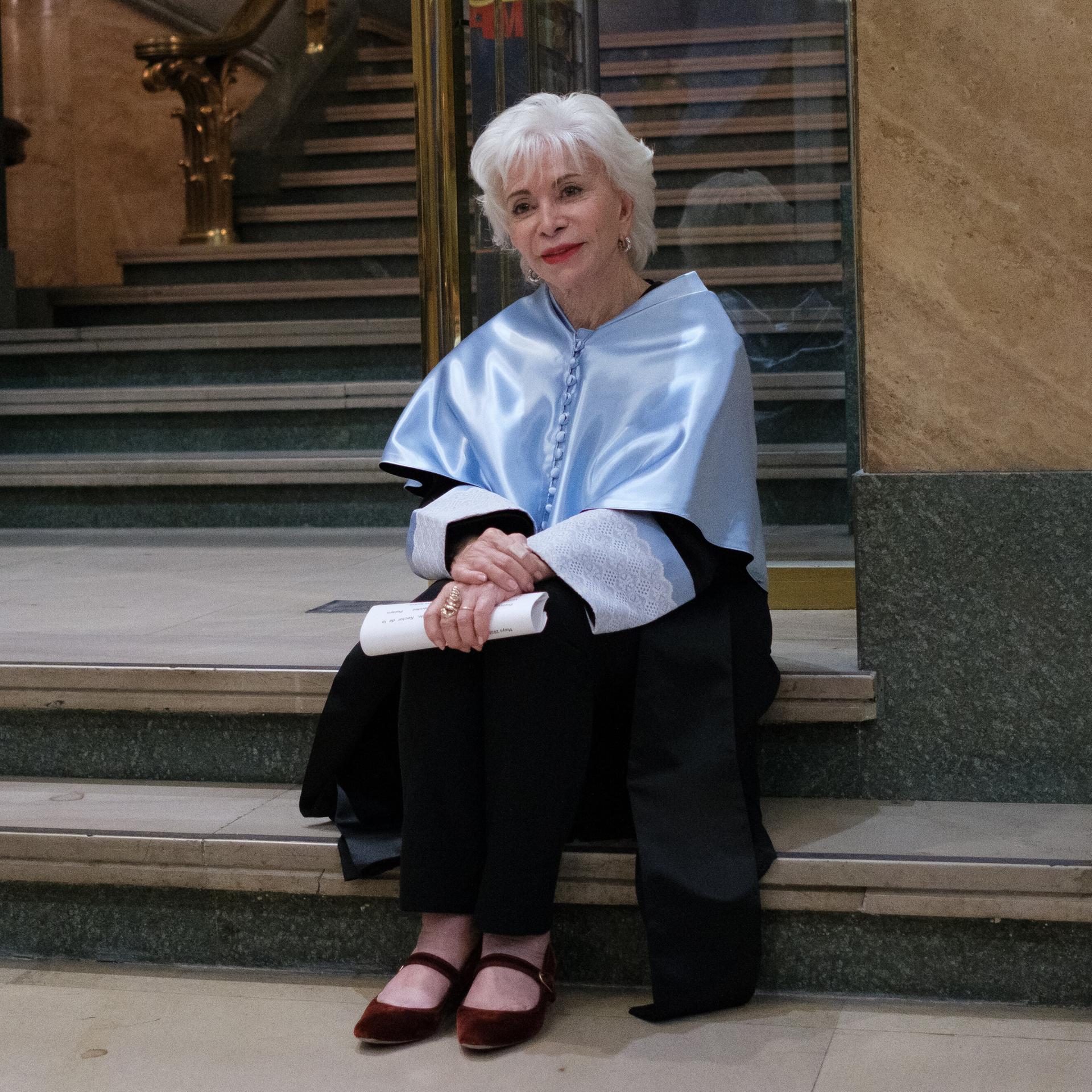 Isabel Allende sitzt auf einer Treppe. Sie hat kurzes, weißes Haar und hält ein gerolltes Stück Papier in der Hand. 
