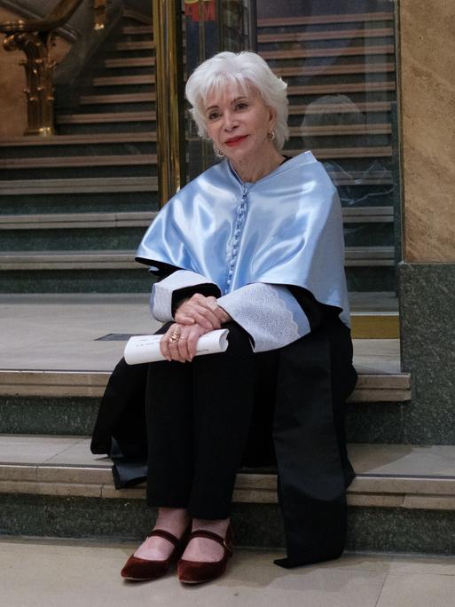 Isabel Allende sitzt auf einer Treppe. Sie hat kurzes, weißes Haar und hält ein gerolltes Stück Papier in der Hand. 