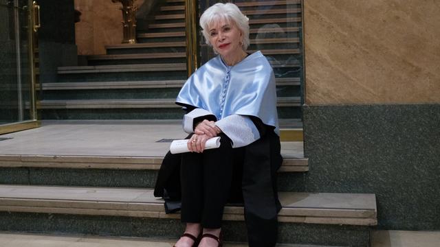 Isabel Allende sitzt auf einer Treppe. Sie hat kurzes, weißes Haar und hält ein gerolltes Stück Papier in der Hand. 