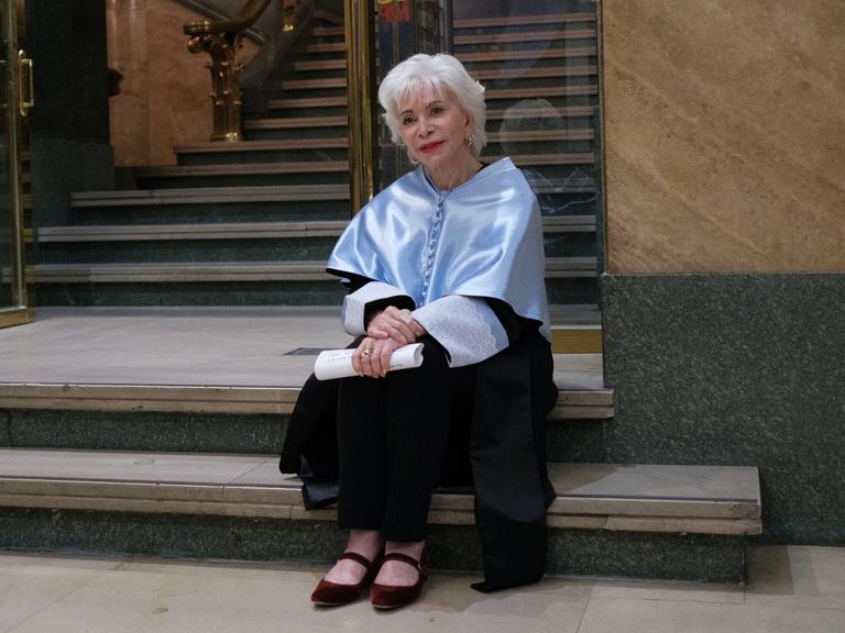Isabel Allende sitzt auf einer Treppe. Sie hat kurzes, weißes Haar und hält ein gerolltes Stück Papier in der Hand. 