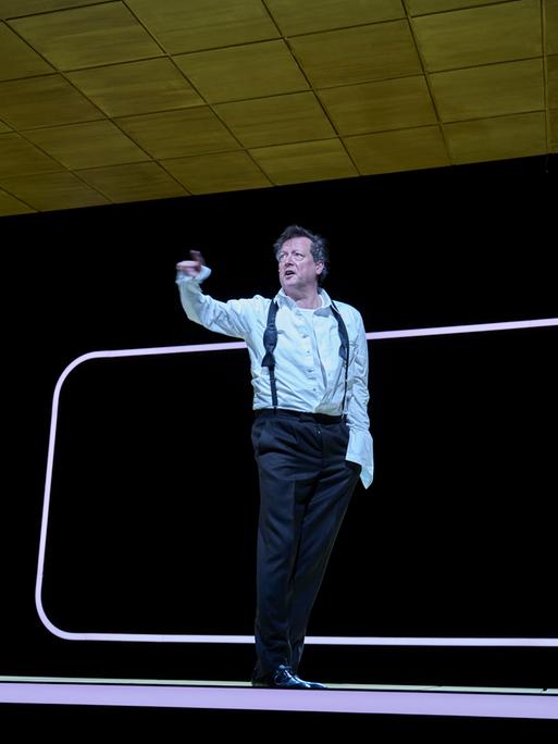 Matthias Brandt steht auf der Bühne mit schlichtem Szenenbild und gestikuliert Richtung Publikum.