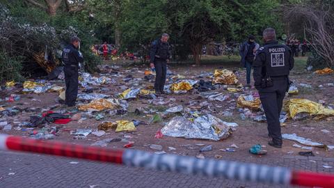 Polizisten stehen hinter einer Absperrung in einem Park. Auf dem Boden liegt Müll, vor allem goldene Rettungsdecken.