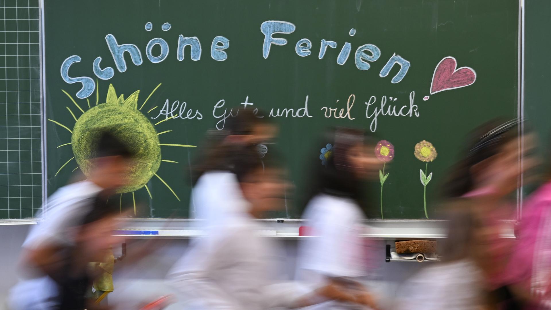 Schulkinder einer Grundschule laufen am letzten Schultag vor den Sommerferien an einer Tafel mit der Aufschrift: "Schöne Ferien! Alles Gute und viel Glück" vorbei.