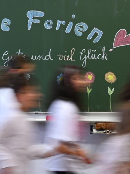 Schulkinder einer Grundschule laufen am letzten Schultag vor den Sommerferien an einer Tafel mit der Aufschrift: "Schöne Ferien! Alles Gute und viel Glück" vorbei.