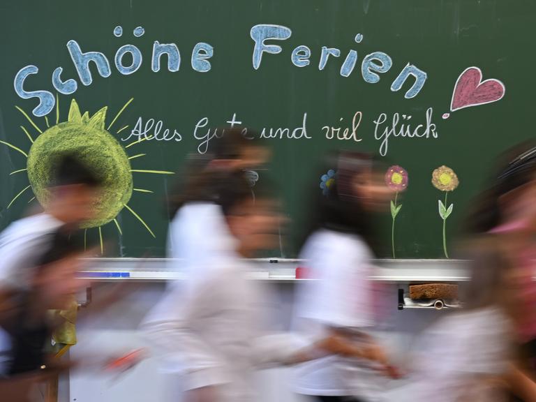 Schulkinder einer Grundschule laufen am letzten Schultag vor den Sommerferien an einer Tafel mit der Aufschrift: "Schöne Ferien! Alles Gute und viel Glück" vorbei.