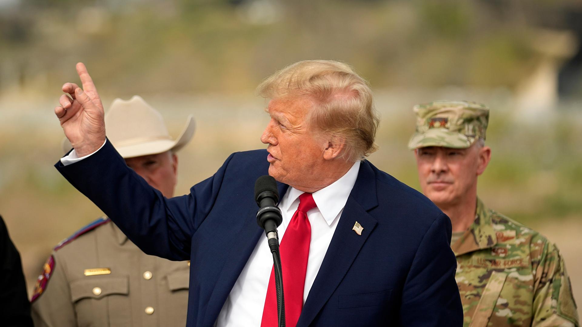 Donald Trump steht an einem Mikrofon und hälte eine Rede. Er hält die rechte Hand erhoben. Hinter ihm stehen ein Soldat und ein Sherrif.