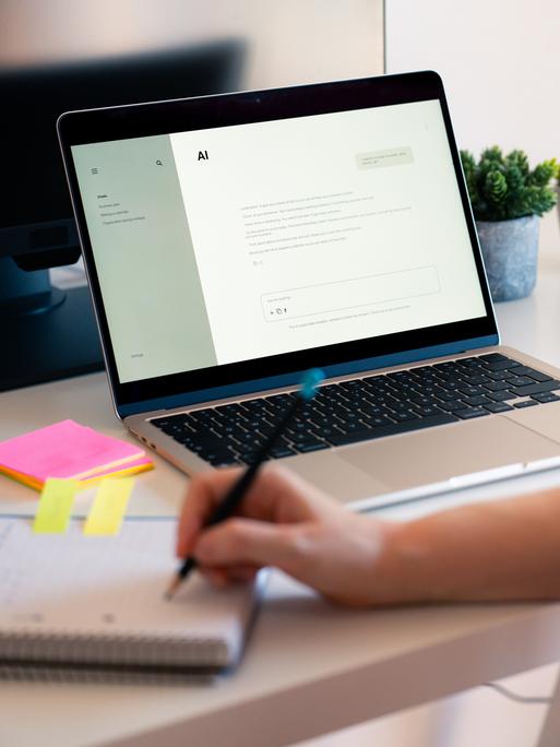 Eine Person hat einen KI-Chatbot auf einem Laptop geöffnet und macht daneben handschriftliche Notizen auf einem Block. Eine Person hat einen KI-Chatbot auf einem Laptop geöffnet und macht daneben handschriftliche Notizen auf einem Block.