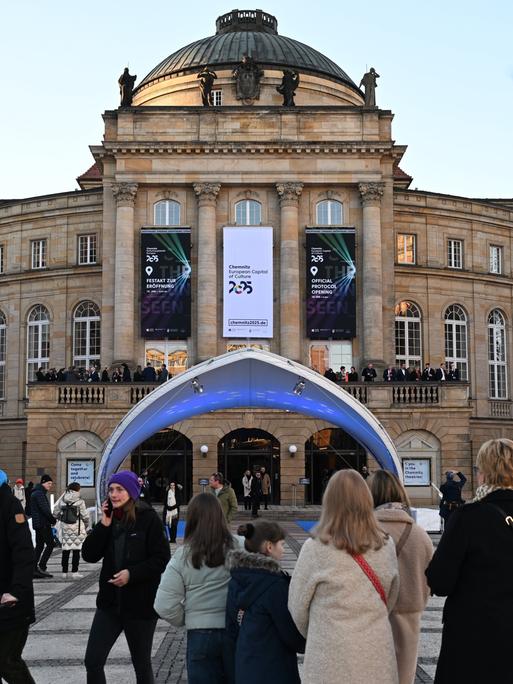 Menschen stehen vor dem Opernhaus in Chemnitz . Dort hängt ein Banner auf dem in englischer Sprache steht: Chemnitz - Kultur-Hauptstadt von europa 2025.