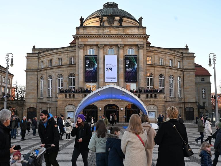 Menschen stehen vor dem Opernhaus in Chemnitz . Dort hängt ein Banner auf dem in englischer Sprache steht: Chemnitz - Kultur-Hauptstadt von europa 2025.