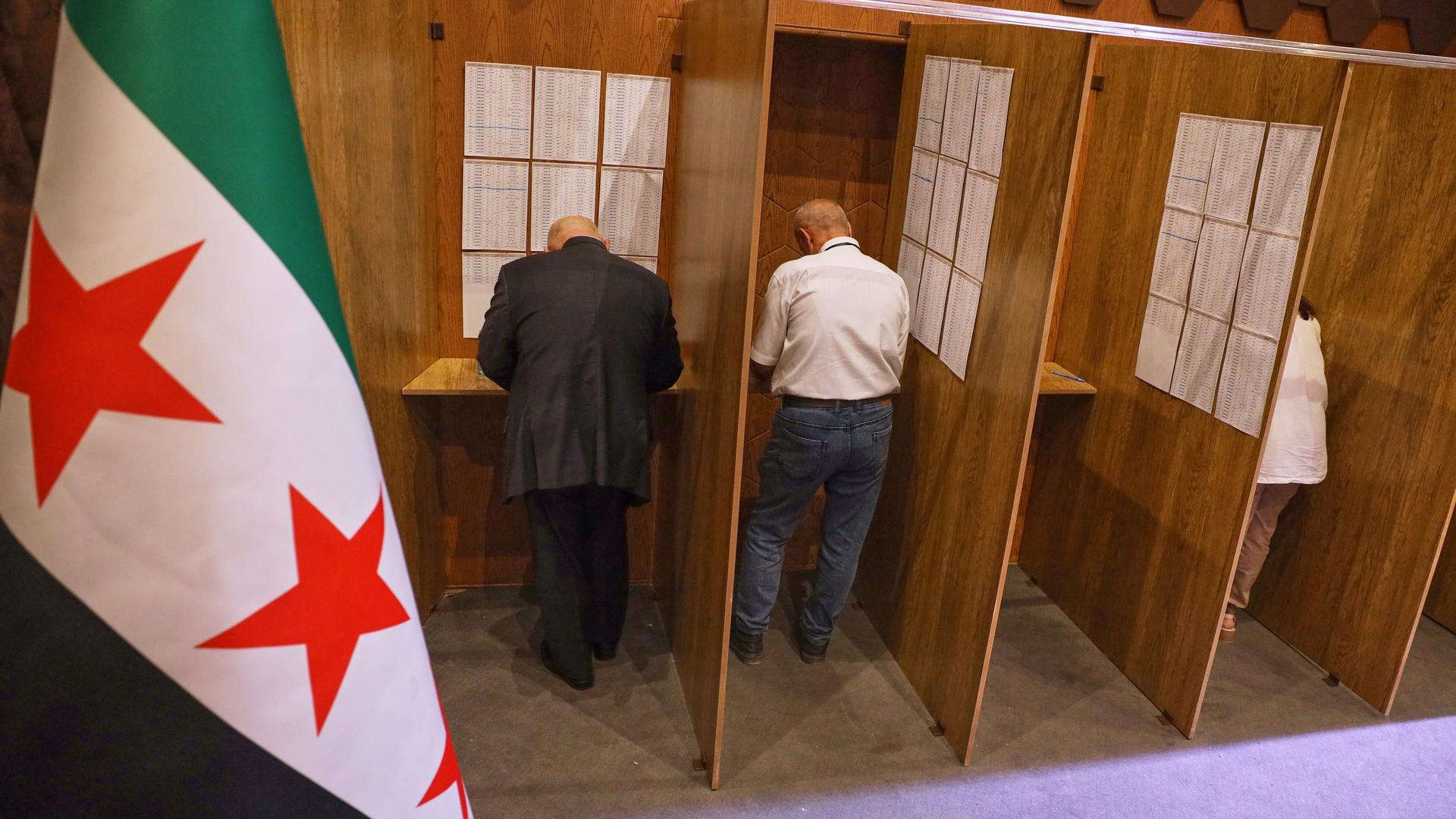 Blick aus höherer Perspektive auf zwei Wahlkabinen, in denen zwei Männer gerade ihre Stimmzettel ausfüllen. Links im Vordergrund eine syrische Nationalflagge. Blick aus höherer Perspektive auf zwei Wahlkabinen, in denen zwei Männer gerade ihre Stimmzettel ausfüllen. Links im Vordergrund eine syrische Nationalflagge.