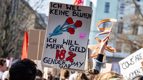 Menschen halten Schilder mit der Aufschrift „Ich will keine Blumen, ich will mehr Geld. 18 % Gender Pay Gap“ auf einer Demonstration in die Luft