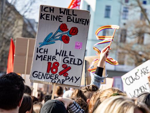 Menschen halten Schilder mit der Aufschrift „Ich will keine Blumen, ich will mehr Geld. 18 % Gender Pay Gap“ auf einer Demonstration in die Luft Menschen halten Schilder mit der Aufschrift „Ich will keine Blumen, ich will mehr Geld. 18 % Gender Pay Gap“ auf einer Demonstration in die Luft