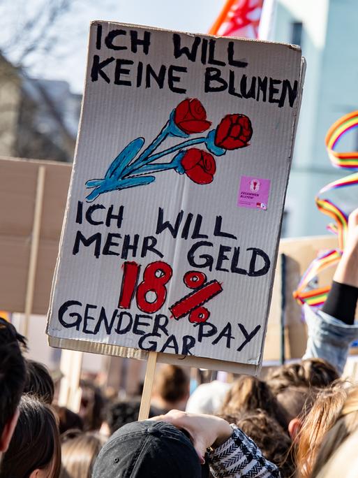 Menschen halten Schilder mit der Aufschrift „Ich will keine Blumen, ich will mehr Geld. 18 % Gender Pay Gap“ auf einer Demonstration in die Luft Menschen halten Schilder mit der Aufschrift „Ich will keine Blumen, ich will mehr Geld. 18 % Gender Pay Gap“ auf einer Demonstration in die Luft