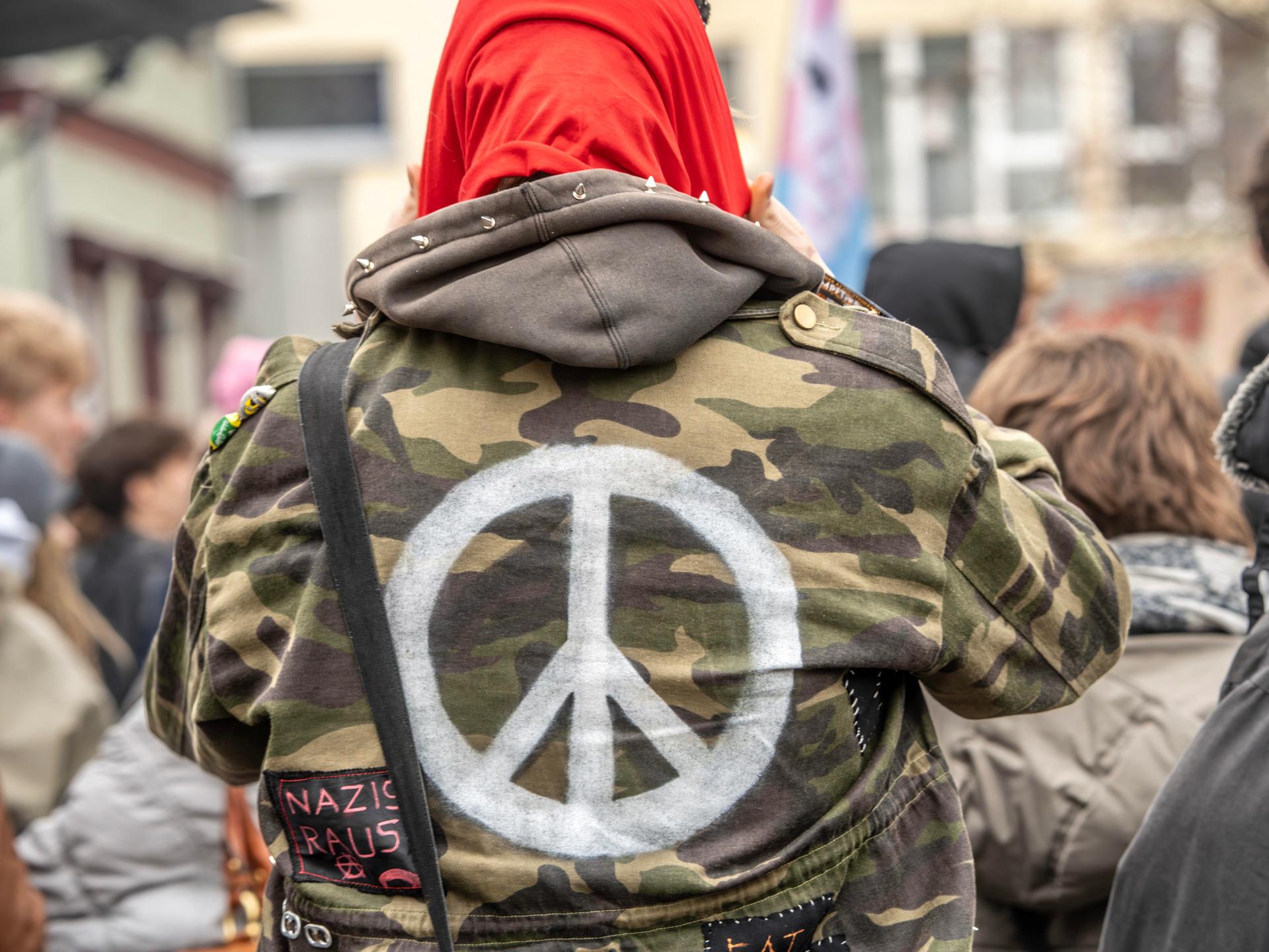 Ein Demonstrant trägt eine Bundeswehrjacke, auf die ein weißes Peace-Zeichen auf dem Rücken aufgemalt ist. 