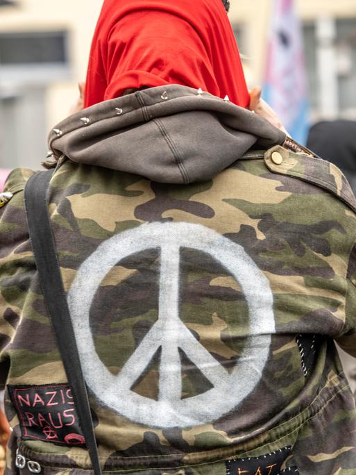 Ein Demonstrant trägt eine Bundeswehrjacke, auf die ein weißes Peace-Zeichen auf dem Rücken aufgemalt ist. 