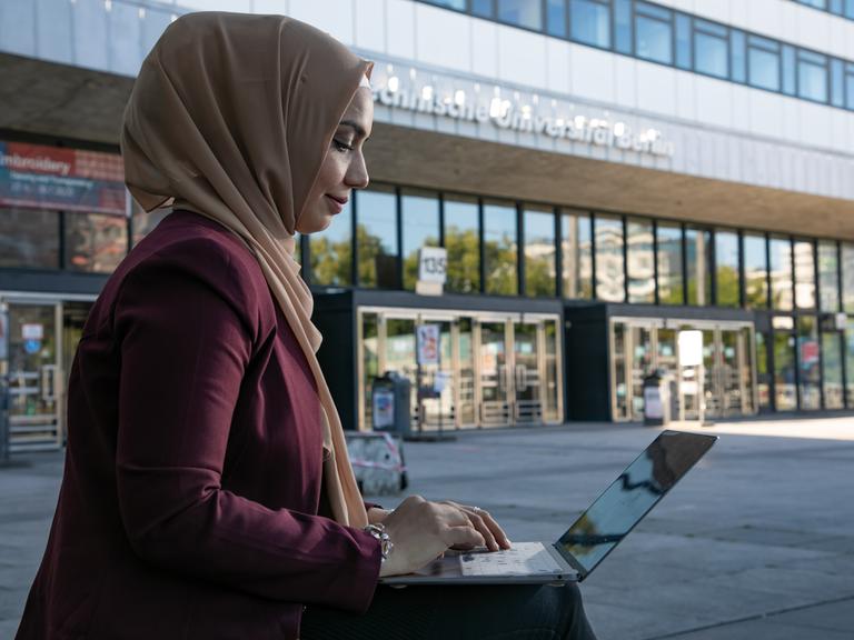 Eine junge Frau mit muslimischer Kopfbedeckung ist im Profil zu sehen. Sie sitzt im Freien vor einem Gebäude der Technischen Universität Berlin  und arbeitet an einem Laptop.