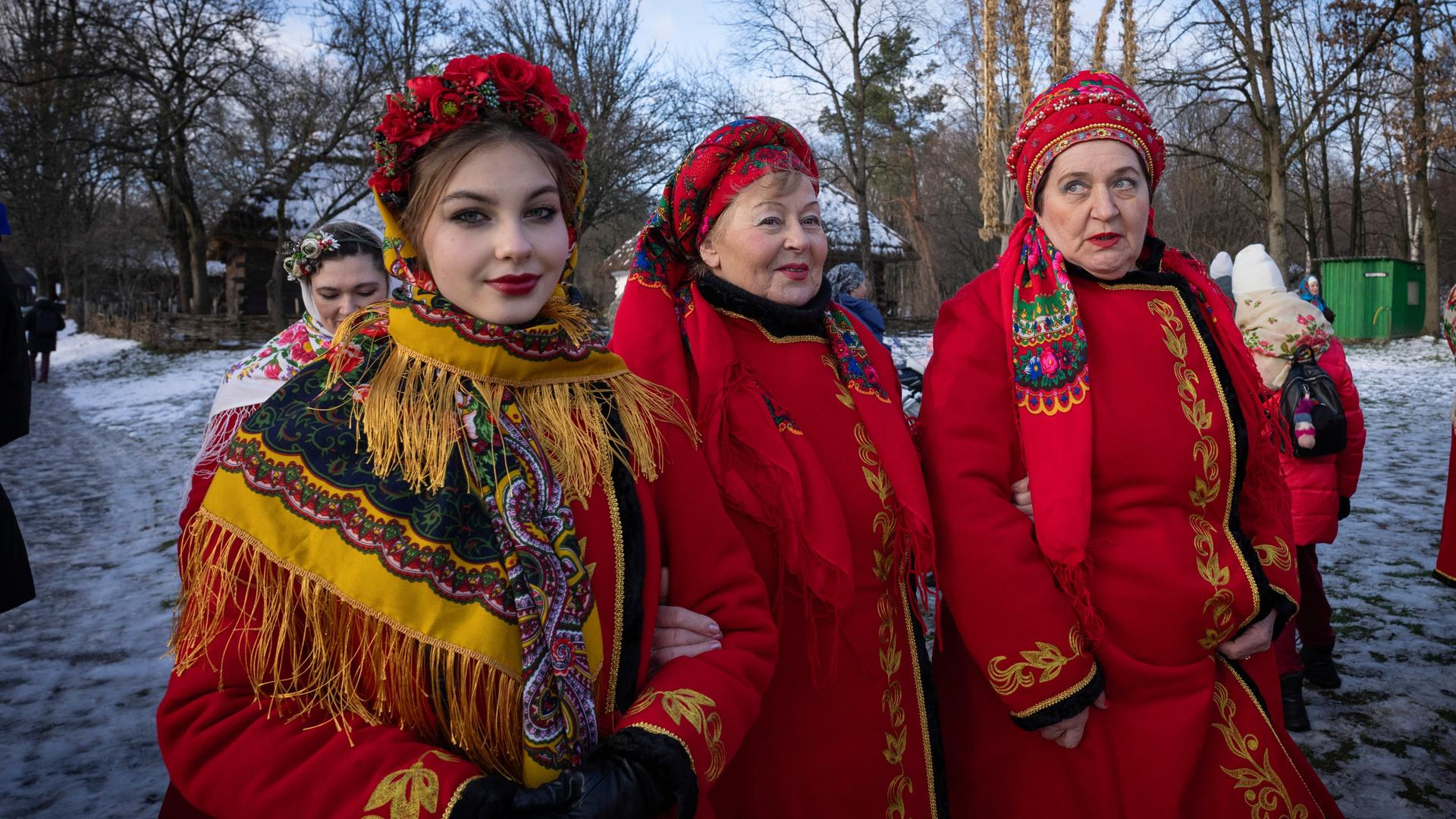 Drei Frauen in traditionellen ukrainischer Kleider mit roten langen Mänteln, die reicht veriehrt sind, gehen über eine schneebedeckte Straße. Szene aus dem Dorf Pirogovo in der Nähe der Hauptstadt Kiew. Aufnahme vom 25.12.2023