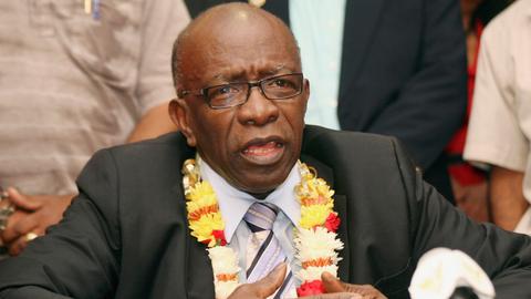 Jack Warner sitzt im Anzug und mit Blumenkranz um den Hals vor einem Mikrofon.