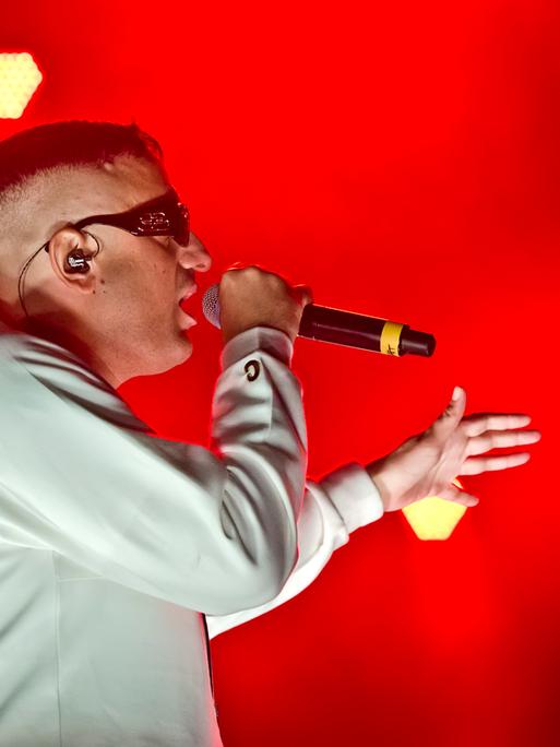 Der Rapper Haftbefehl in einer weissen Jacke mit einer Sonnenbrille bei einem Auftritt auf einer rot erleuchteten Bühne.