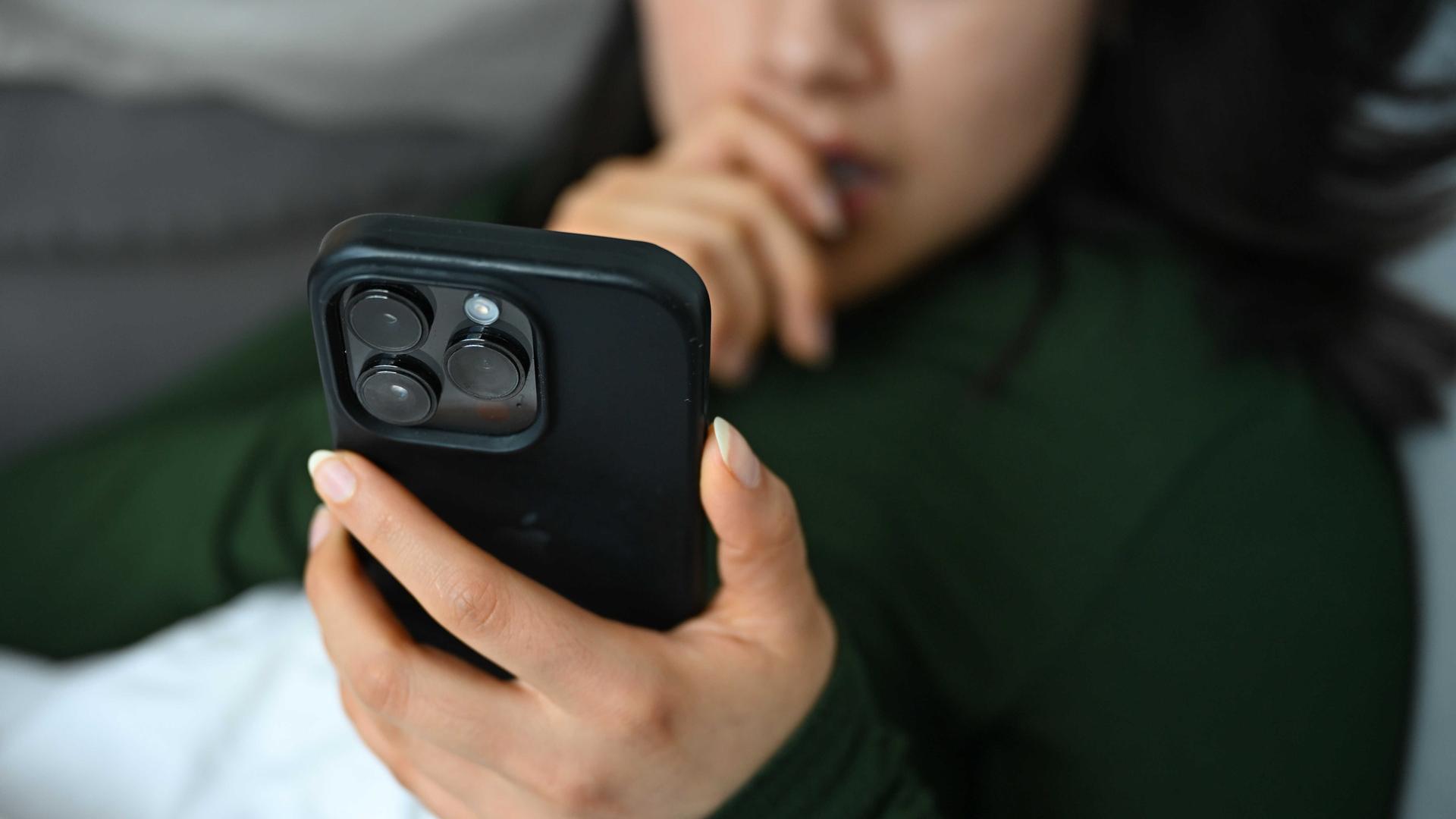 Eine junge Frau liegt mit ihrem Handy auf der Couch und blickt auf das Display.