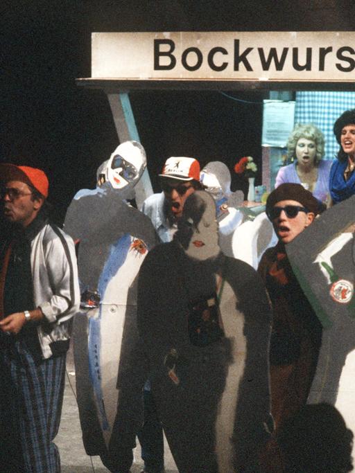 Szene des Rock-Musicals "Linie 1" im Berliner Grips-Theater an einem Bockwurststand, vor dem Menschen als Touristen verkleidet mit Sonnenbrille und Fotoapparaten sowie zusätzlichen Pappmaché-Figuren singen.