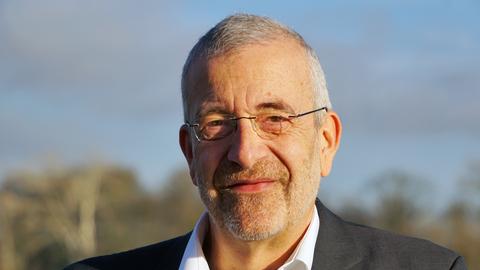 Porträt von Reinhard Küchler: Ein Mann mit ergrautem Haar und einem Drei-Tage-Bart steht draußen vor einem unscharfen Hintergrund, trägt eine randlose Brille, Hemd und dunkles Sakko.Er blickt in Richtung Kamera und hat ein Lächeln angedeutet.