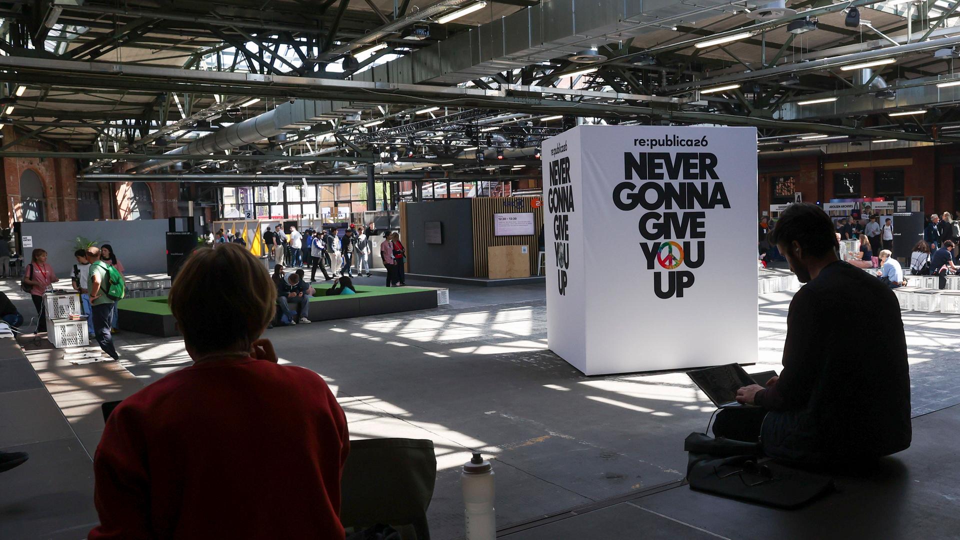 Das Motto steht auf einem weißen Würfel in einer Messehalle. Drumherum sitzen und stehen Besucher. 