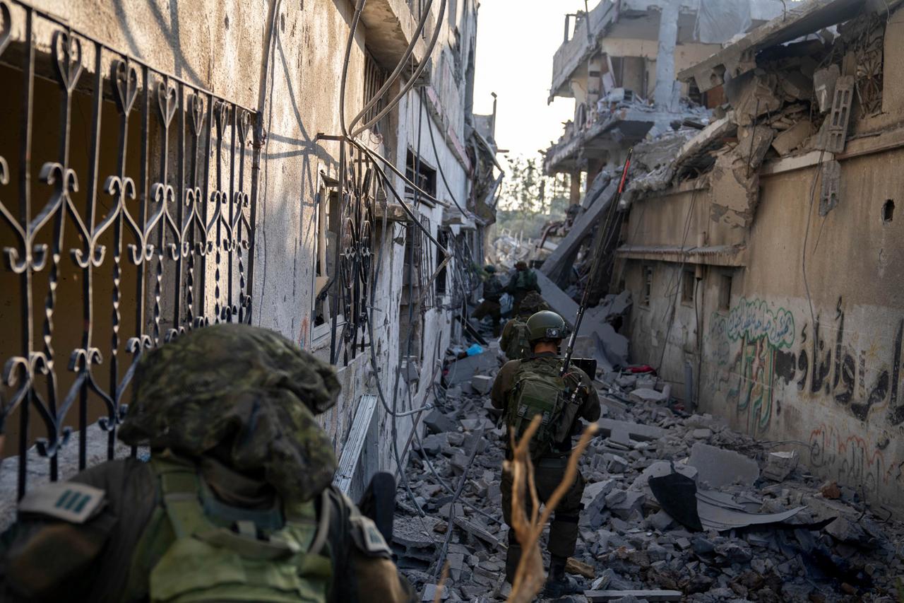 Mehrere Soldaten gehen im Gänsemarsch durch eine schmale Gasse, die mit Trümmern von Häusern übersät ist.