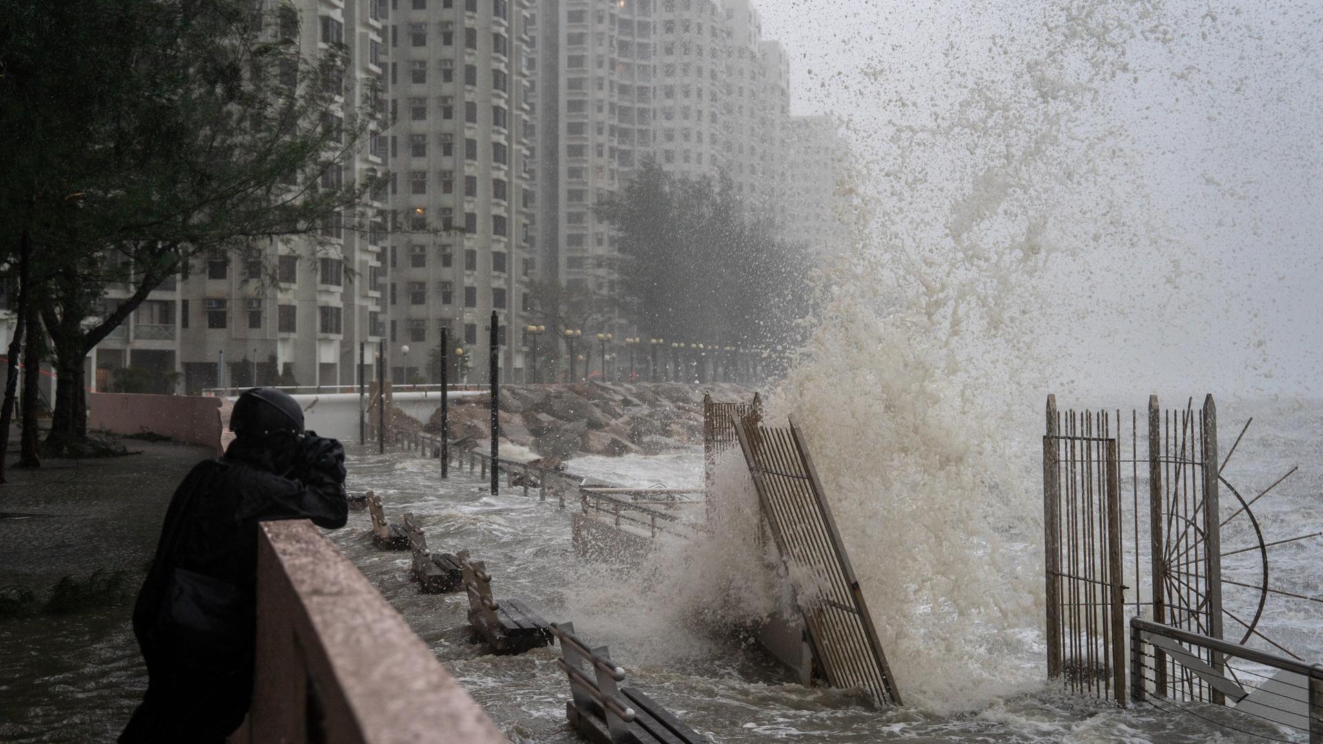 Ein Mann fotografiert hohe Wellen in Hongkong. Im Hintergrund sind Hoch-Häuser.