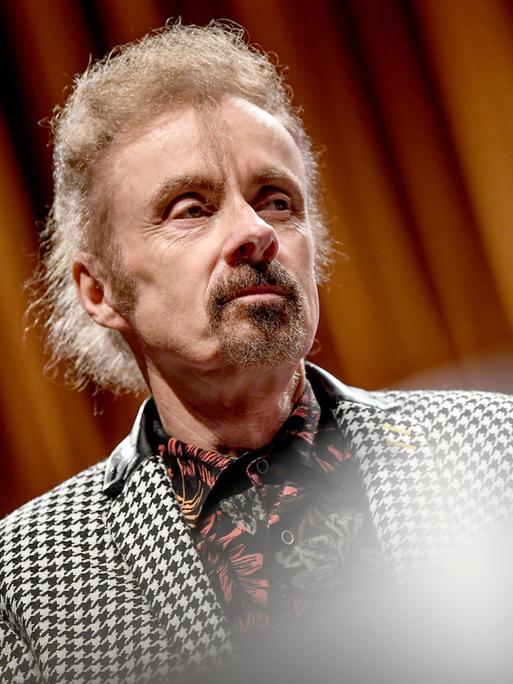 Porträt von T.C. Boyle. Er steht bei einer Veranstaltung auf einer Bühne vor einem Vorhang und trägt einen Blazer mit schwarzweißem Hahnentrittmuster.