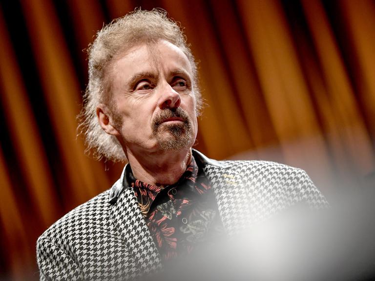 Porträt von T.C. Boyle. Er steht bei einer Veranstaltung auf einer Bühne vor einem Vorhang und trägt einen Blazer mit schwarzweißem Hahnentrittmuster.