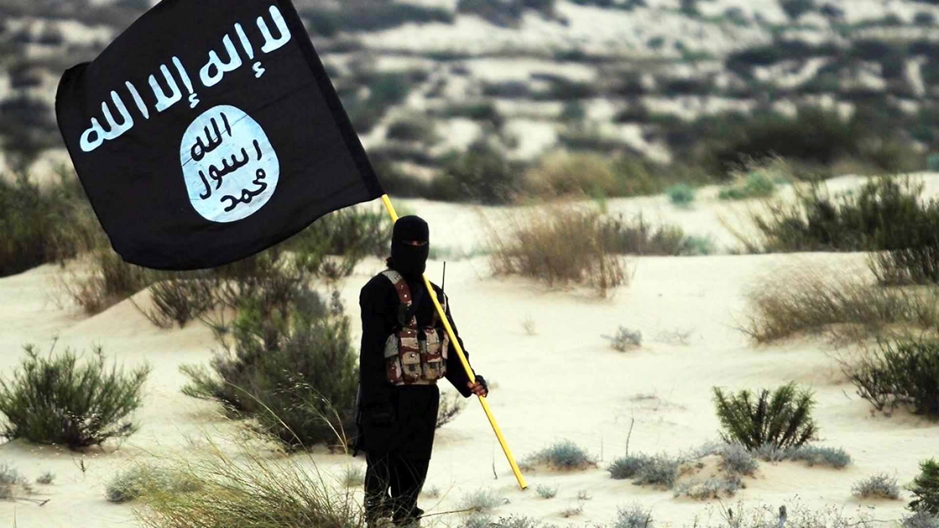 Ein maskierter Kämpfer des Islamischen Staats steht in einer Wüstenlandschaft und schwenkt die schwarze Fahne des IS.