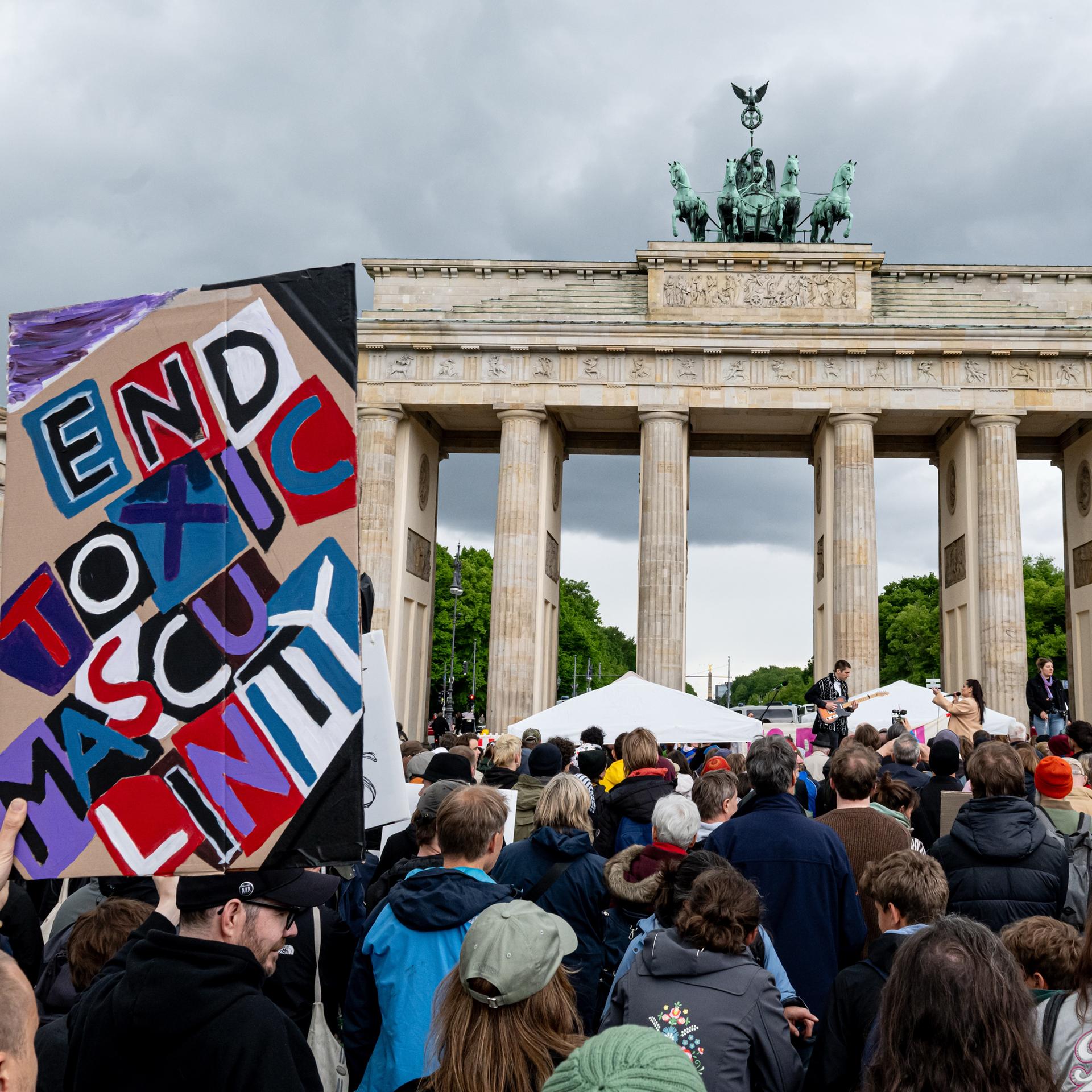 Menschen nehmen an der Demonstration "Männer gegen Gewalt"  teil. Im Vordergrund ein Schild mit der Aufschrift "end toxic masculinity", im Huintergrund das Brandenburger Tor. 