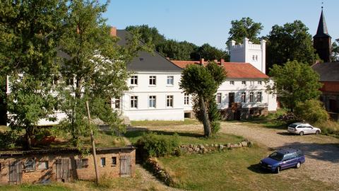 Aufnahme von Schloss Bröllin, im Vordergrund stehen links einige Bäume, das Gebäude mitsamt des mächtigen Turmes an der rechten Seite ist weiß angemalert. Im Hintergrund des Schlosses sind Baumkronen zu erkennen.