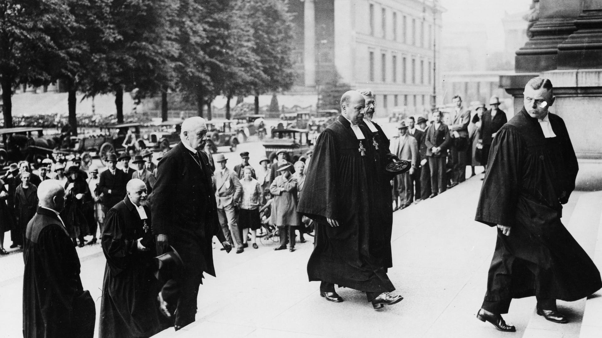 Reichspräsident Paul von Hindenburg schreitet mit evangelischen Pfarrern im Talar die Treppen zum Berliner Dom hoch. 