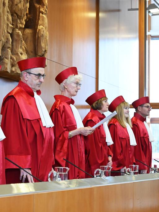 Die Richterinnen und Richter stehen nebeneinander. Sie haben rote Kleidung an. Das ist typisch für die Richter an diesem Gericht. Eine Richterin hält ein Papier in der Hand. Sie liest etwas vor.  Die Richterinnen und Richter stehen nebeneinander. Sie haben rote Kleidung an. Das ist typisch für die Richter an diesem Gericht. Eine Richterin hält ein Papier in der Hand. Sie liest etwas vor.