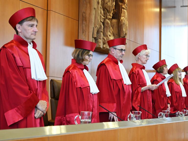Die Richterinnen und Richter stehen nebeneinander. Sie haben rote Kleidung an. Das ist typisch für die Richter an diesem Gericht. Eine Richterin hält ein Papier in der Hand. Sie liest etwas vor.  Die Richterinnen und Richter stehen nebeneinander. Sie haben rote Kleidung an. Das ist typisch für die Richter an diesem Gericht. Eine Richterin hält ein Papier in der Hand. Sie liest etwas vor.