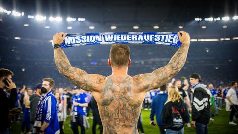 Ein Schalke Fan feiert den Aufstieg des FC Schalke 04 nach dem Spiel gegen St. Pauli am 07.05.2022. Er ist von hinten zu sehen und reckt einen Schal in zwischen beiden Armen in die Luft. Darauf steht: "Mission Wiederaufstieg".
