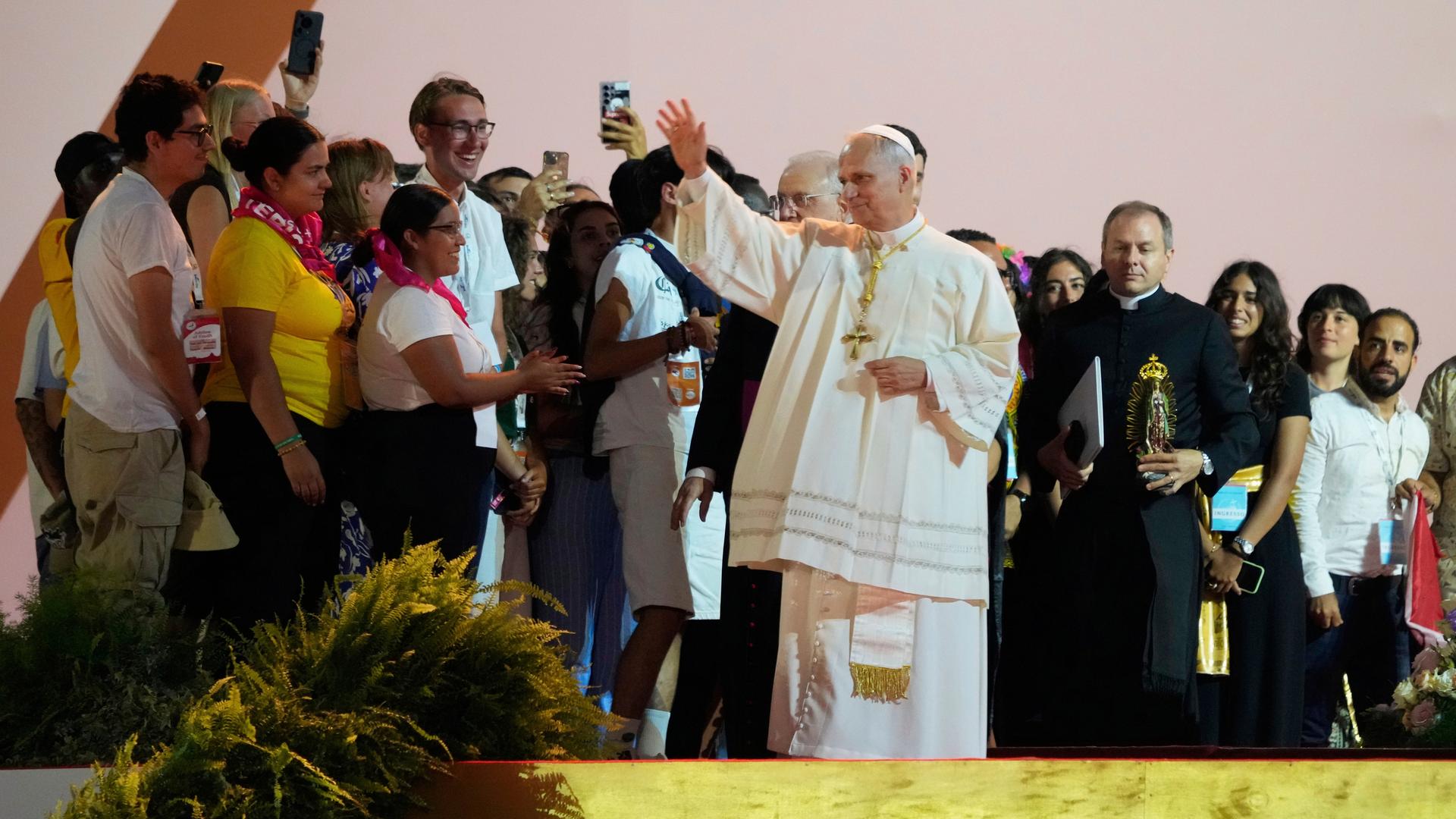 Der Papst im weißen Ornat steht in einem Zelt vor einer großen Gruppe junger Menschen und hebt die rechte Hand. Viele der Jugendlichen lächeln, manche filmen mit ihren Handys.