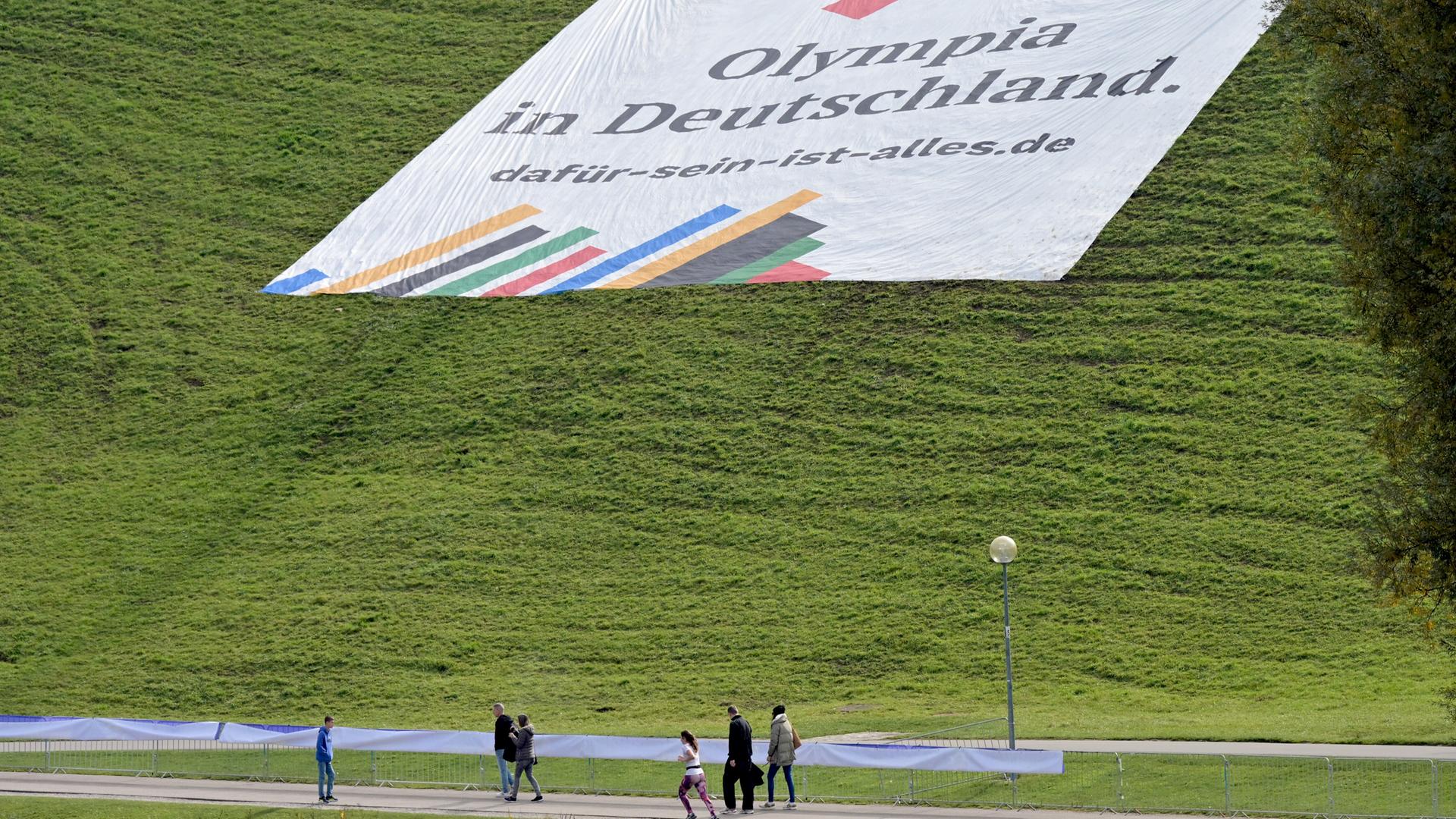 Ein Plakat mit der Aufschrift „"Olympia in Deutschland. dafür-sein-ist-alles.de"“ liegt auf dem Olympiaberg der Stadt München. Spaziergänger gehen daran vorbei.