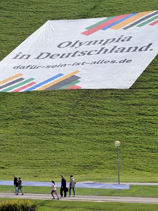 Ein Plakat mit der Aufschrift „"Olympia in Deutschland. dafür-sein-ist-alles.de"“ liegt auf dem Olympiaberg der Stadt München. Spaziergänger gehen daran vorbei.