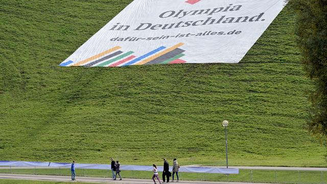 Ein Plakat mit der Aufschrift „"Olympia in Deutschland. dafür-sein-ist-alles.de"“ liegt auf dem Olympiaberg der Stadt München. Spaziergänger gehen daran vorbei.