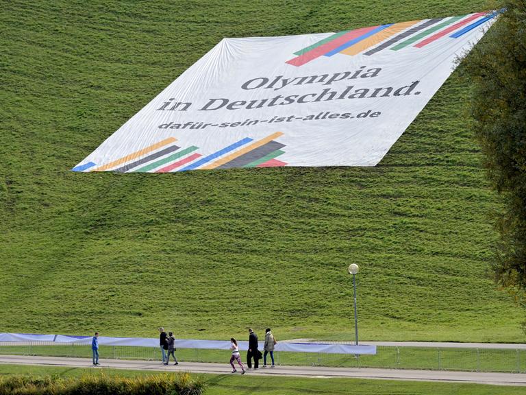 Ein Plakat mit der Aufschrift „"Olympia in Deutschland. dafür-sein-ist-alles.de"“ liegt auf dem Olympiaberg der Stadt München. Spaziergänger gehen daran vorbei.