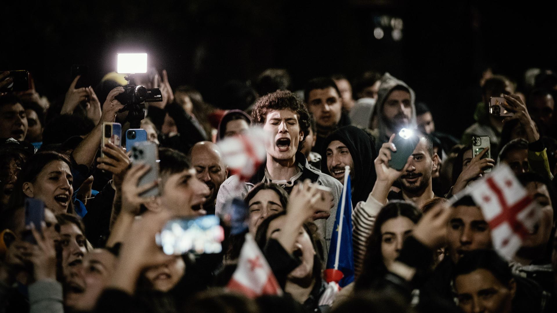 Demonstranten stehen vor dem Parlament in Tiflis. Sie halten georgische Fahnen und Handys mit eingeschalteten Lictern in den Händen. Sie schreien.