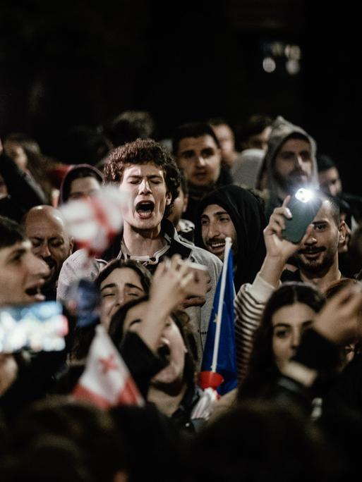 Demonstranten stehen vor dem Parlament in Tiflis. Sie halten georgische Fahnen und Handys mit eingeschalteten Lictern in den Händen. Sie schreien.