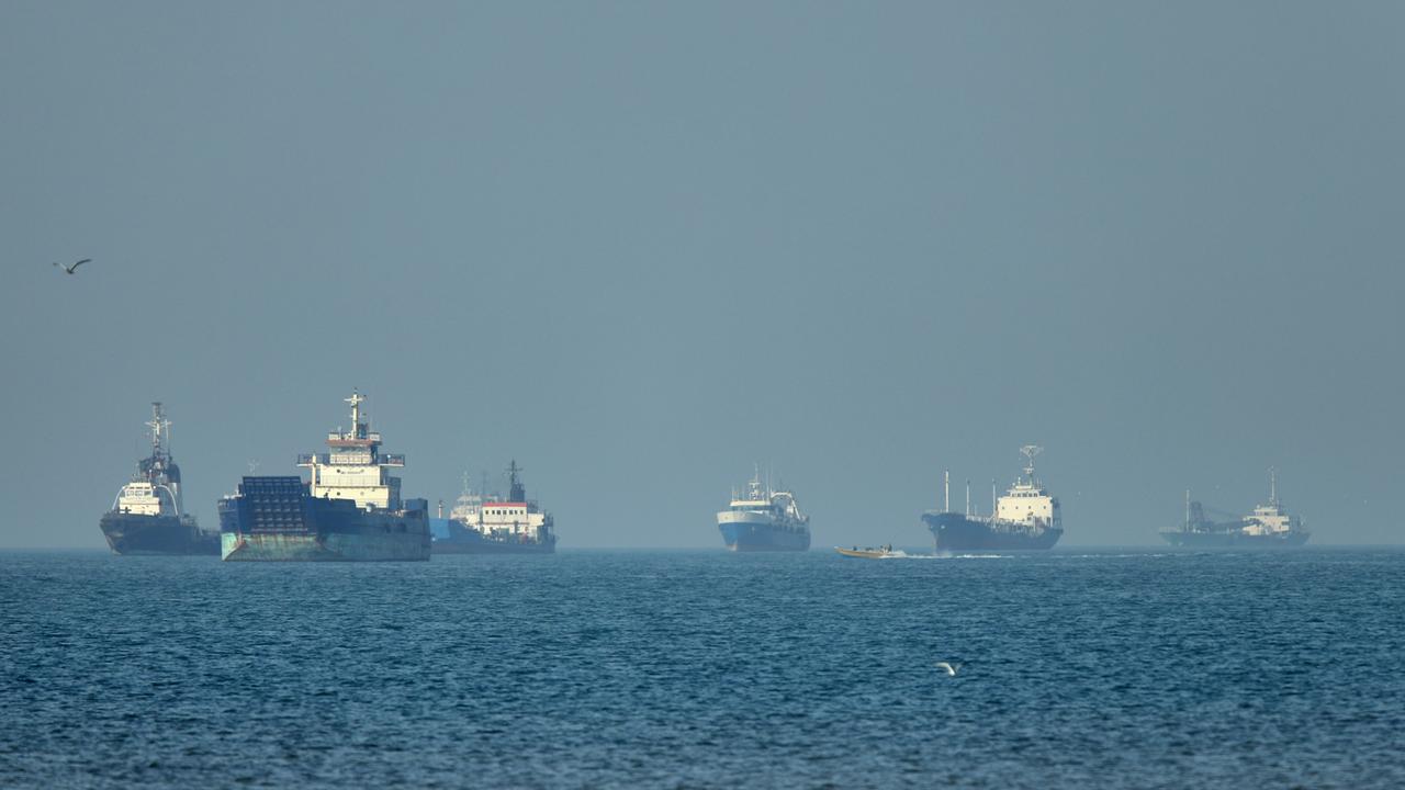 Frachtschiffe, darunter Massengutfrachter und Stückgutschiffe, liegen vor der Küste vor Anker, während im Vordergrund ein kleines Motorboot vorbeifährt 