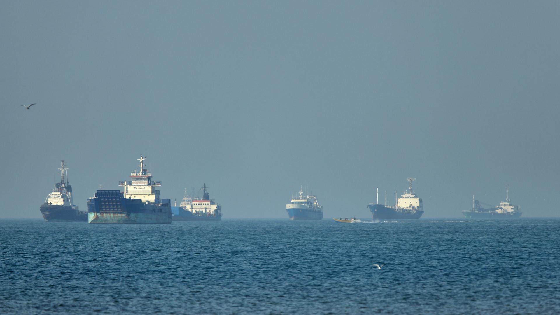 Frachtschiffe, darunter Massengutfrachter und Stückgutschiffe, liegen vor der Küste vor Anker, während im Vordergrund ein kleines Motorboot vorbeifährt 